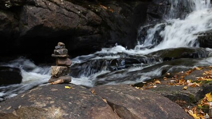 Poster - Stationary low view at stack of rocks cairn on edge of cascading river with fall leaves