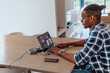 © .shock - African American man in glasses sitting at a table in a modern living room, using a laptop for business video chat, conversation with friends and entertainment