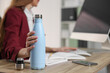 © New Africa - Woman holding thermos bottle at workplace, closeup. Space for text