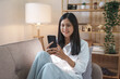 © amnaj - Beautiful Asian woman sitting on sofa in living room using mobile phone playing games and various entertainment programs at home holiday and having fun on vacation.