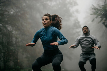 Wall Mural - Two people in sports outfits have an active HIIT workout in the forest in the morning during a rainy day. A woman and a man jumping 