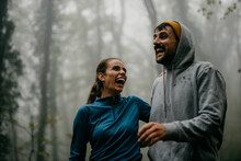 Cheerful And Tired Couple Walking In The Foggy Forest After Jogging During A Rainy Day. Fit Woman And Man Embracing And Smiling After A Running.