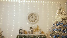 Christmas And New Year 2023 In Ukraine. Decorated Fir-tree With Blue Bulbs And Golden Yellow Bows. Fireplace, Golden Reindeer Toys And Clock On Wall