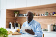 © PhotoAlto - Senior African-American man sitting in kitchen eating fruit for breakfast while checking messages on digital tablet