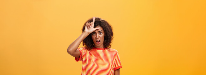 Girl thinks she loser. Portrait of gloomy bothered and displeased african american woman with afro hairstyle showing l word over forehead complaining feeling gloomy and unhappy over orange background