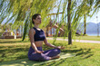 Young beautiful woman practicing meditation outdoors. Yogini sitting on green mat and meditating in the park with the sea view. Close up, copy space, background.