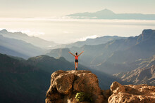 Young Girl Standing On A Rock With The Amazing View Of Roque Nublo Natural Park And Mount Teide In The Background, Gran Canary, Canary Islands, Spain