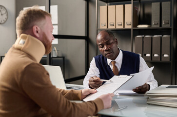 Portrait of black insurance broker helping man with neck brace filling legal forms after accident