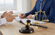 © Studio Romantic - Wooden gavel on sound block and golden scales of justice on office desk, with attorney meeting client in background. Cropped close up closeup. Law, justice, lawyer services, legal consultation concept
