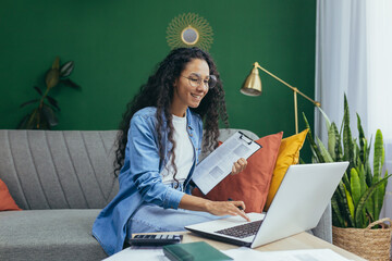 Wall Mural - Happy hispanic woman working remotely with documents at home, paperwork woman using calculator and laptop sitting on sofa in living room.