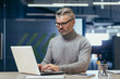 © Liubomir - Portrait of a senior gray-haired man, a businessman, director, founder, who is concentrating on working in the office at a desk with a laptop.