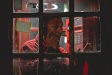 Portrait Of Young And Attractive Model Guy With Red Jacket Smoking A Cigarette In A Red Phone Booth In The City In The Night
