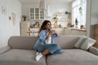 © DimaBerlin - Independent woman holding cup of coffee sits on sofa at home, dreaming, looking into distance enjoying loneliness. Optimistic Italian lady dressed in casual style contemplates future of weekend