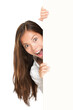 Sign people - woman peeking over billboard paper poster. Excited woman looking surprised. Beautiful brunette with long hair. Asian Caucasian female model isolated cutout PNG on transparent background