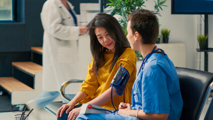 Wall Mural - Female nurse doing cardiology consultation with asian patient in waiting area lobby, measuring hypertension with tonometer. Checking blood and pulse pressure with medical instrument.