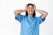 © Cookie Studio - Portrait of carefree happy woman holding hands on head with disbelief and excitement, close eyes and smiling, standing in blue t-shirt over white background