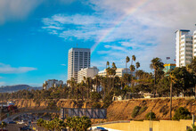 White Hotels And Office Buildings In The City Skyline Surrounded By Tall Lush Green Palm Trees And Pants With Parked Cars And Blue Sky, Clouds And A Rainbow In Santa Monica California USA