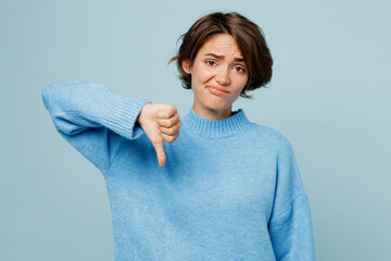 Wall Mural - Young sad unhappy caucasian woman wearing knitted sweater look camera showing thumb down dislike gesture isolated on plain pastel light blue cyan background studio portrait. People lifestyle concept.