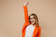 © ViDi Studio - Young fun employee business woman corporate lawyer wear classic formal orange suit glasses work in office do winner gesture celebrate clenching fists raise up hand isolated on plain beige background.
