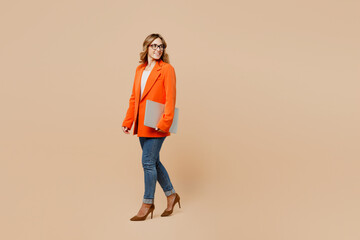 Full body young employee IT business woman corporate lawyer wear classic formal orange suit glasses work in office hold closed laptop pc computer look aside on area isolated on plain beige background.