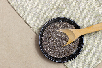 Wall Mural - Healthy food, Chia seeds with wooden spoon in black bowl on linen cloth over cardboard background