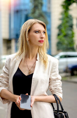 Wall Mural - Portrait with an attractive blonde business lady in an oatmeal jacket and a black dress with an iPhone in her hand on the street. Summer urban landscape - Background. Women. Business. concept