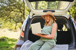 Senior happy woman working with laptop from the car trunk or boot in camper sunny summer park - digital nomad freedom lifestyle concept, travel freelancer and vacations of retired old age people.