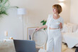 © nazariykarkhut - Happy smiling young woman taking a coffee break as she stands at her laptop computer working from a home office in stylish white livingroom.
