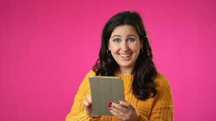 Portrait of happy smiling young woman 20s in sweater shopping on tablet computer point with finger isolated on pink background studio. 