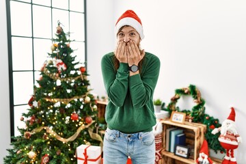 Wall Mural - Redhead man with long beard wearing christmas hat by christmas tree laughing and embarrassed giggle covering mouth with hands, gossip and scandal concept