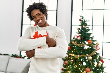 Wall Mural - Young african american man holding gift standing by christmas tree at home.