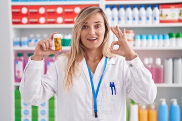 Sticker - Young blonde woman working at pharmacy drugstore holding pills doing ok sign with fingers, smiling friendly gesturing excellent symbol