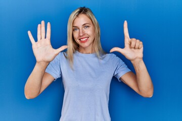 Poster - Beautiful blonde woman wearing casual t shirt over blue background showing and pointing up with fingers number seven while smiling confident and happy.