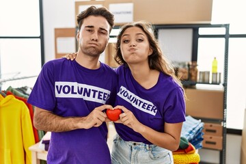 Wall Mural - Young hispanic couple wearing volunteer t shirt holding heart puffing cheeks with funny face. mouth inflated with air, catching air.