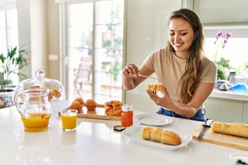 Poster - Young beautiful hispanic woman preparing breakfast putting jam on bread at the kitchen