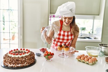 Young beautiful hispanic woman smiling confident holding tray with cupcakes at the kitchen