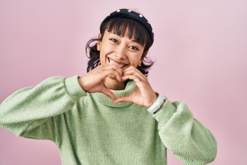 Poster - Young beautiful woman standing over pink background smiling in love doing heart symbol shape with hands. romantic concept.