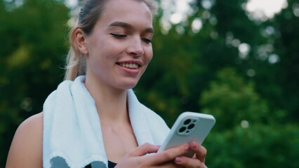 Caucasian female runner carrying tower on her neck using smartphone, texting, scrolling apps, walking in the park after training. Sport, people and communication concept.