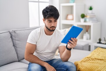 Wall Mural - Hispanic man with beard using touchpad sitting on the sofa relaxed with serious expression on face. simple and natural looking at the camera.