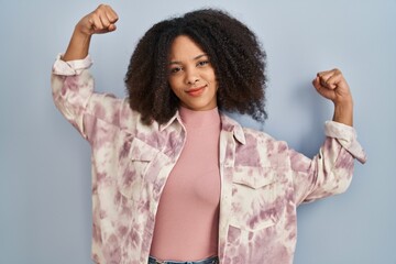 Sticker - Young african american woman standing over blue background showing arms muscles smiling proud. fitness concept.