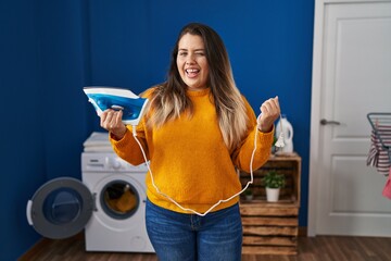 Canvas Print - Young hispanic woman holding electric iron winking looking at the camera with sexy expression, cheerful and happy face.