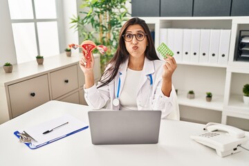 Canvas Print - Young hispanic doctor woman holding anatomical female genital organ and birth control pills puffing cheeks with funny face. mouth inflated with air, catching air.