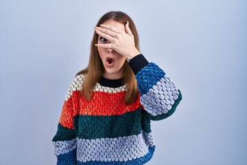 Poster - Young hispanic girl standing over blue background peeking in shock covering face and eyes with hand, looking through fingers with embarrassed expression.