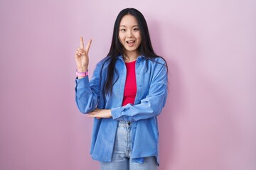 Sticker - Young asian woman standing over pink background smiling with happy face winking at the camera doing victory sign with fingers. number two.