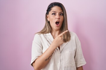 Wall Mural - Blonde woman standing over pink background surprised pointing with finger to the side, open mouth amazed expression.