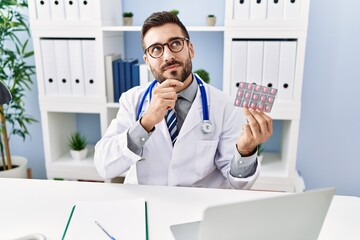 Wall Mural - Handsome hispanic man wearing doctor uniform holding prescription pills serious face thinking about question with hand on chin, thoughtful about confusing idea