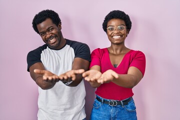 Poster - Young african american couple standing over pink background smiling with hands palms together receiving or giving gesture. hold and protection