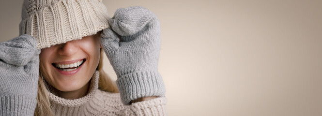 smiling woman wearing warm clothes hiding face under knitted wool hat on beige background. trendy winter fashion clothing. banner with copy space