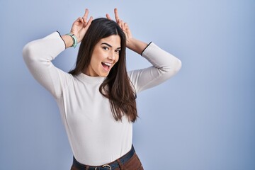 Poster - Young brunette woman standing over blue background posing funny and crazy with fingers on head as bunny ears, smiling cheerful