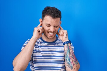 Poster - Young hispanic man standing over blue background covering ears with fingers with annoyed expression for the noise of loud music. deaf concept.